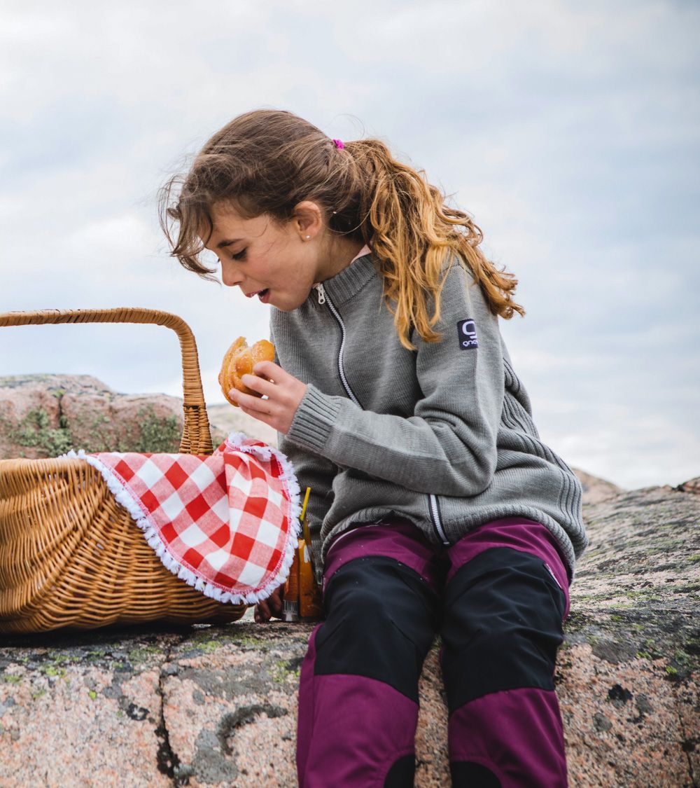 Barn som sitter på en klippa och äter en bulle, iklädd ulltröja och friluftsbyxor från Gneis