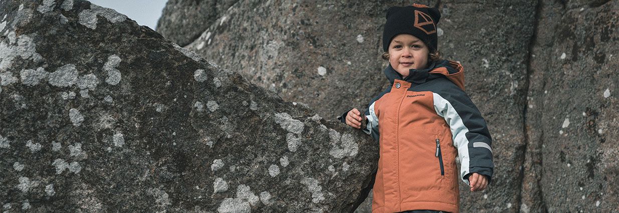Ett barn/ junior som står bredvid en bergsvägg med en varm svart, vit orange jacka ifrån Didirksson