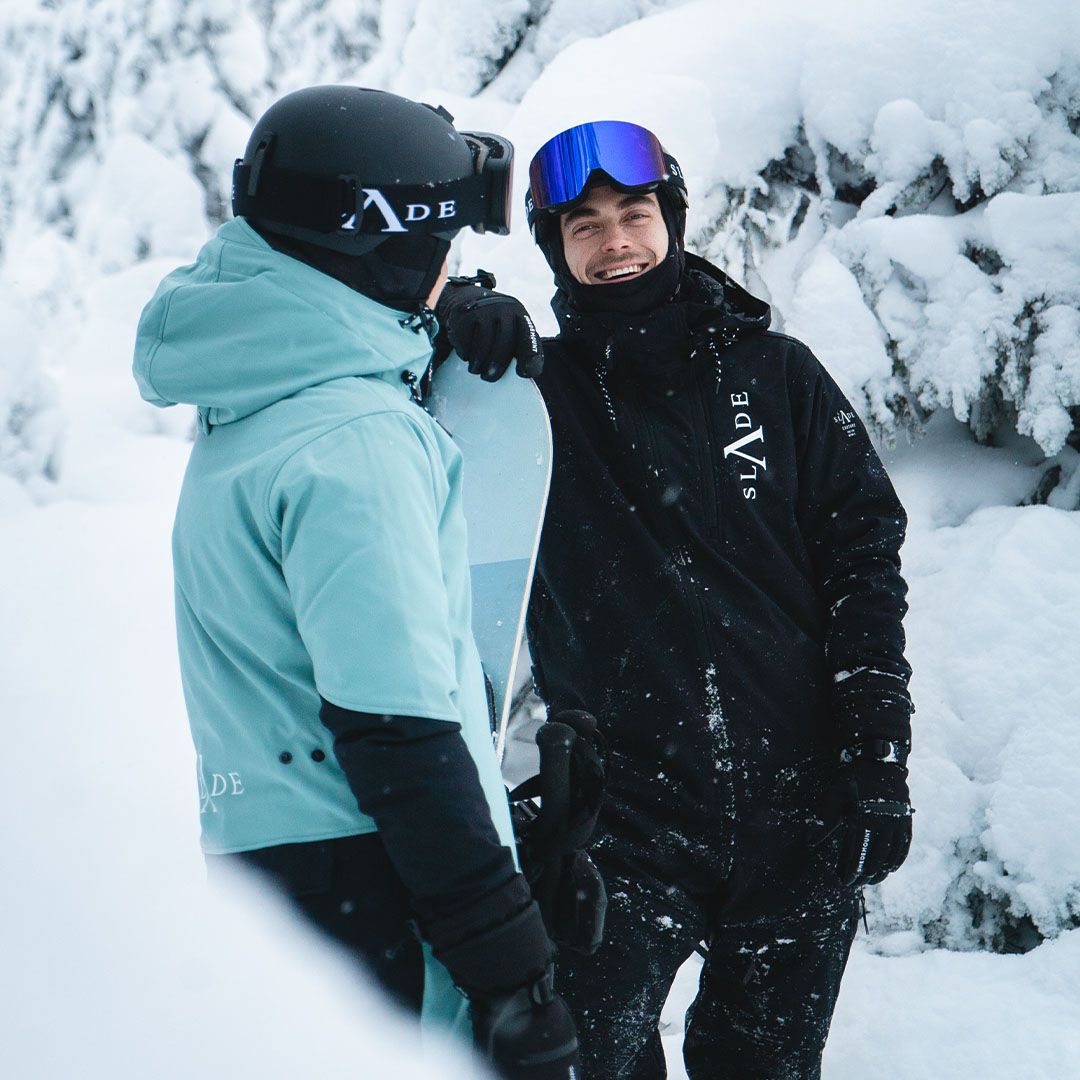 Zwei Menschen tragen Skikleidung in den Farben türkis und schwarz, Helm und Skibrille von Slade in einer Winterlandschaft.
