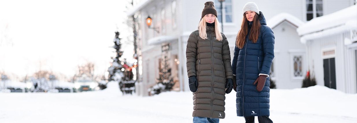 Två tjejer som står utomhus i snön med varma långa vinterjackor ifrån Swedemount kollektionen Östersund i färgerna grönt och navy blått