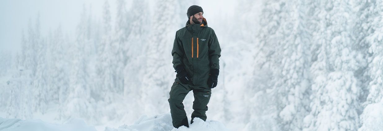 En kille står i snön med en grön Skisuit med orange detaljer ifrån Slade